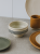 Kremowa ceramiczna miska na podwyższeniu z czarnymi, poziomymi paskami stoi na szarym blacie. Minimalistyczna aranżacja naczyń obejmuje talerze w kolorach ziemi oraz ochrowy kubek w tle.