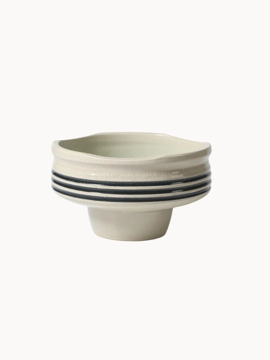 Ceramiczna miska na podwyższeniu w jasnokremowym kolorze, pokryta spękanym szkliwem z trzema czarnymi paskami wokół środka.