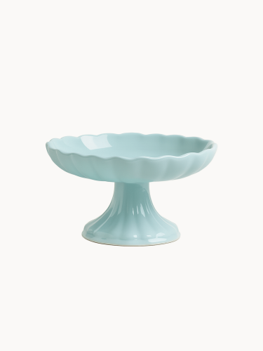 Pastelowa jasnoniebieska ceramiczna patera o stabilnej nóżce i pofalowanej czarce na jednolitym białym tle.